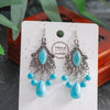 Turquoise and silver earrings on a white card with 'Handmade' text, against a blurred natural background.