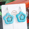 Turquoise star-shaped earrings on a white card with 'Handmade' text, against a blurred natural background.