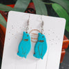 Turquoise cat-shaped earrings on a white card with 'Handmade' text, against a blurred green plant background.