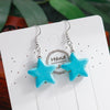 Turquoise star-shaped earrings on a white card with 'Handmade' text, against a blurred green leafy background.