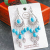 Pair of blue beaded earrings with silver feather charms on a white card, against a dark background.