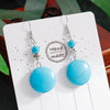 Turquoise earrings on a white card with 'Handmade' label, against a blurred natural background.